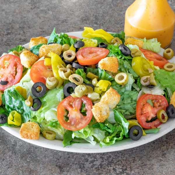 A fresh garden salad with lettuce, tomatoes, olives, pepperoncini, and croutons, with dressing on the side.