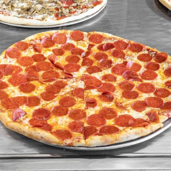 A large, round pepperoni pizza with melted cheese on a metal tray, with another pizza in the background.